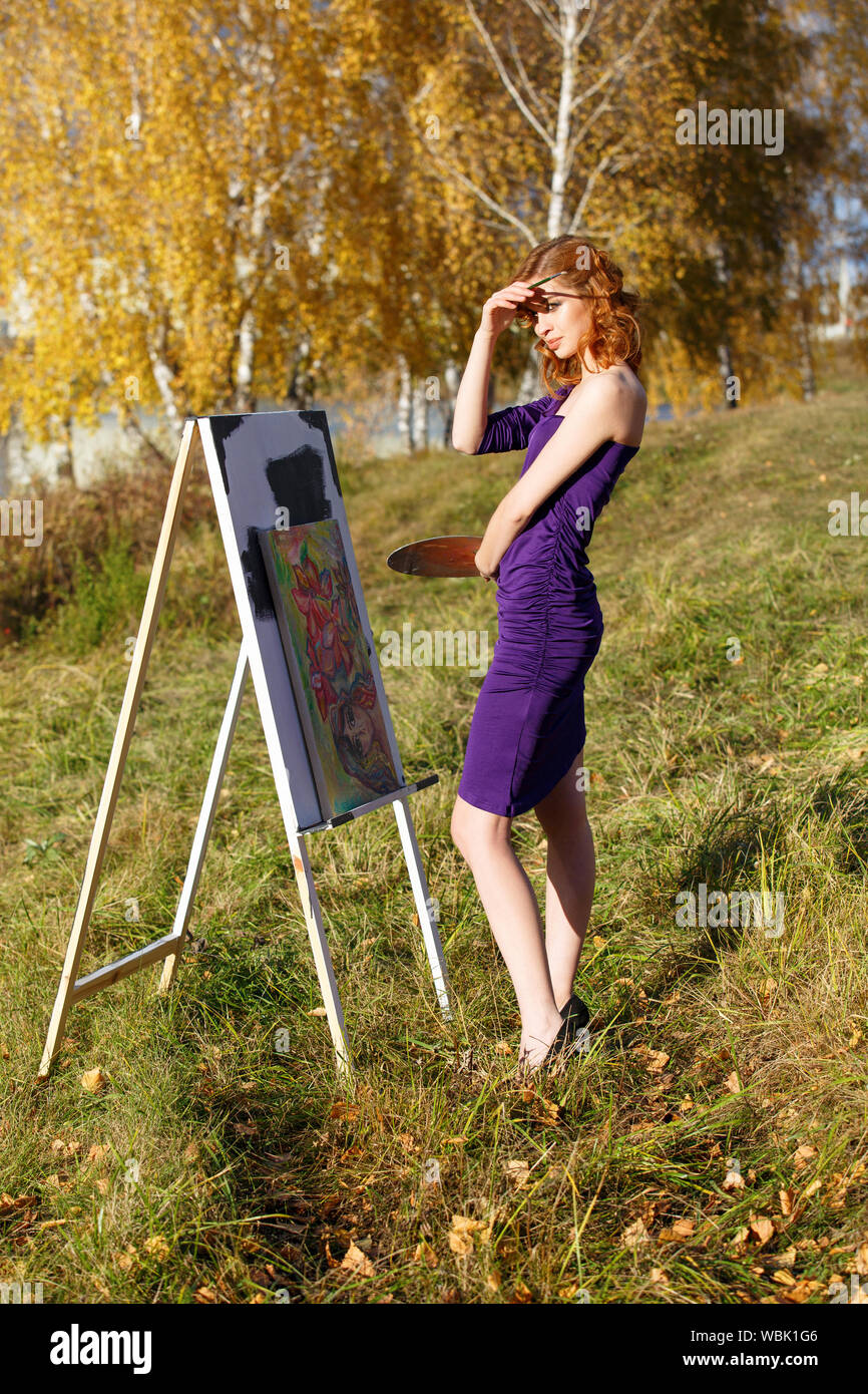 Junge schöne Frau in sexy lila Kleid mit Palette und Pinsel malen im Freien im Herbst Park. Stockfoto