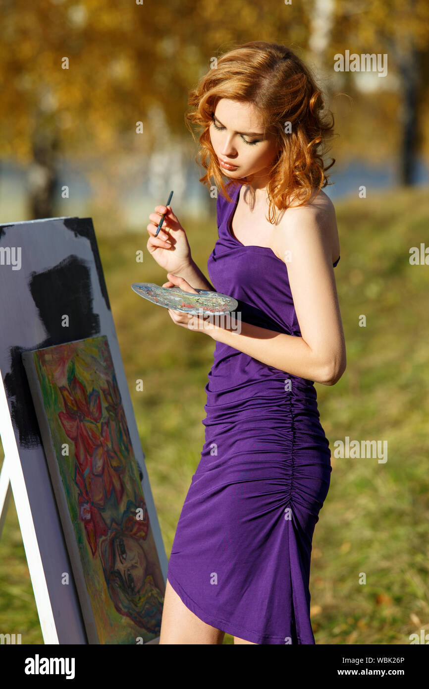 Junge schöne Frau in sexy lila Kleid mit Palette und Pinsel malen im Freien im Herbst Park. Stockfoto