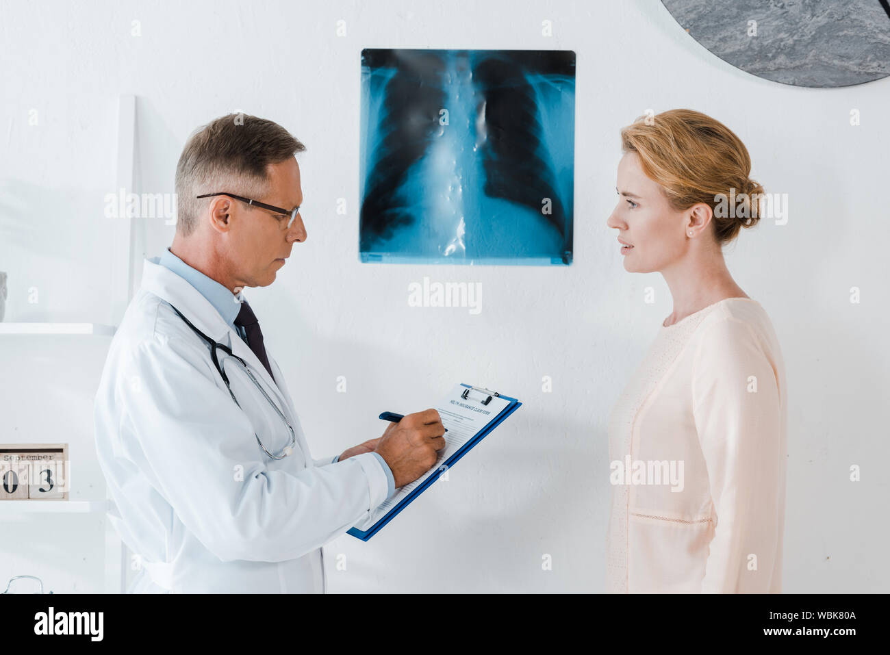 Arzt in Gläsern schreiben Diagnosen über die Zwischenablage in der Nähe von x-ray und die Frau in der Klinik Stockfoto