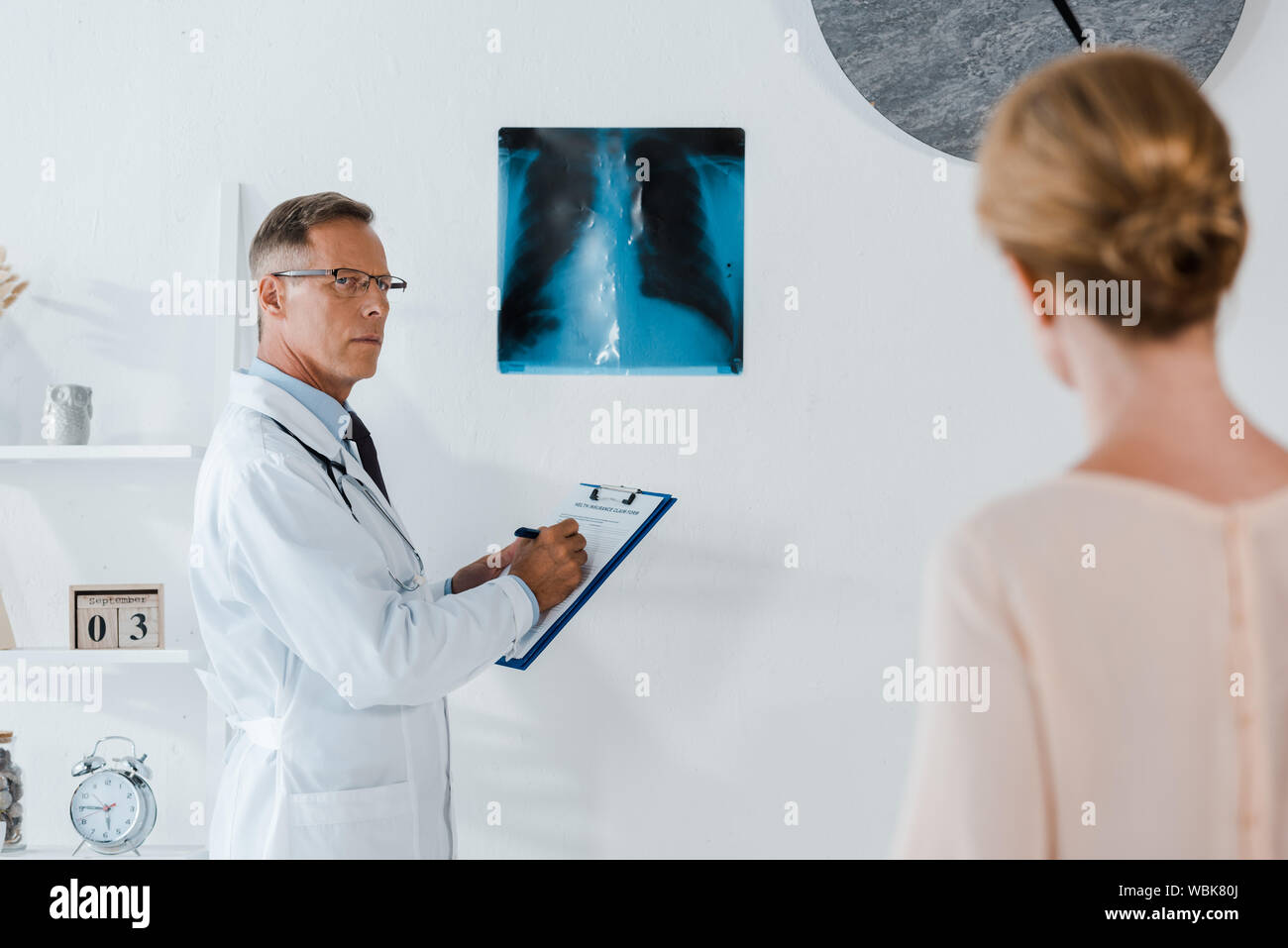 Selektiver Fokus der Arzt in Gläsern schreiben Diagnosen über die Zwischenablage in der Nähe von x-ray und Frau in Klinik Stockfoto