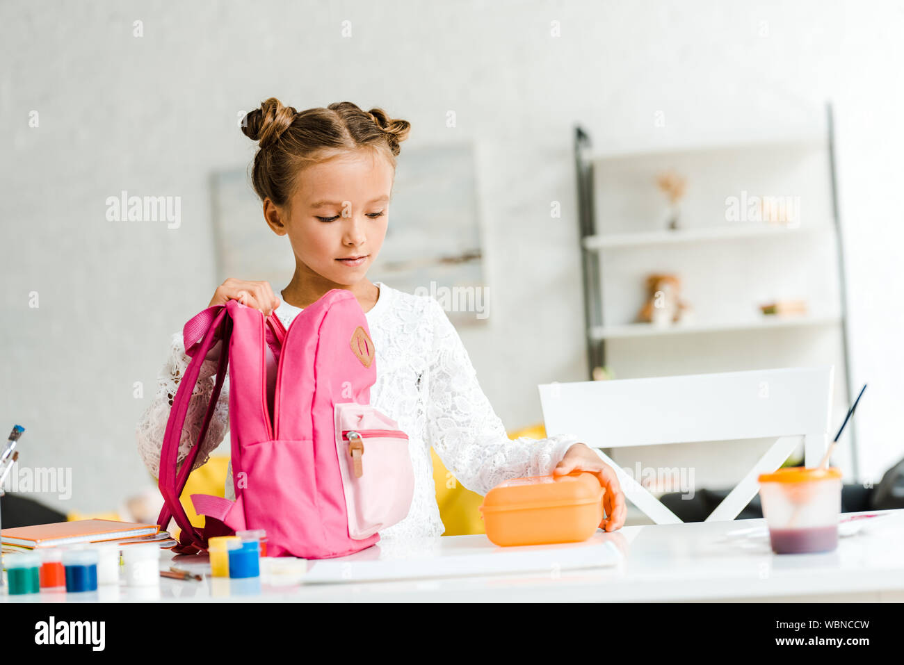 Süßes Schulmädchen holding Lunch Box in der Nähe von Rosa Rucksack Stockfoto