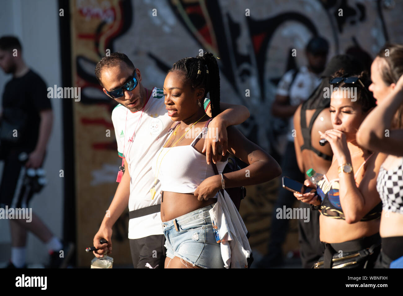 Ein Mann in reflektierender Sonnenbrille mit seinen Arm um eine junge Frau an der Notting Hill Carnival, wie Sie beide Weiß tragen. Stockfoto