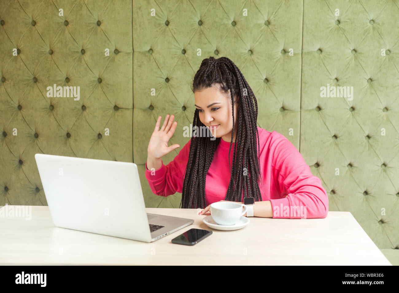 Hallo. Sympathische junge Frau mit schwarzen dreadlocks Frisur in rosa Bluse sitzt und Gruß Arbeitnehmer durch eine Webcam zum Laptop und winkte ihr Stockfoto