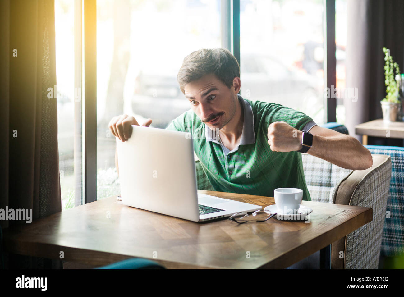 Junge wütend Geschäftsmann in grünem t-shirt Sitzen mit aggressiven Gesicht und Boxing Fäuste und zu seinem Laptop Stanzen versuchen. Problem auf Geschäft und freelan Stockfoto