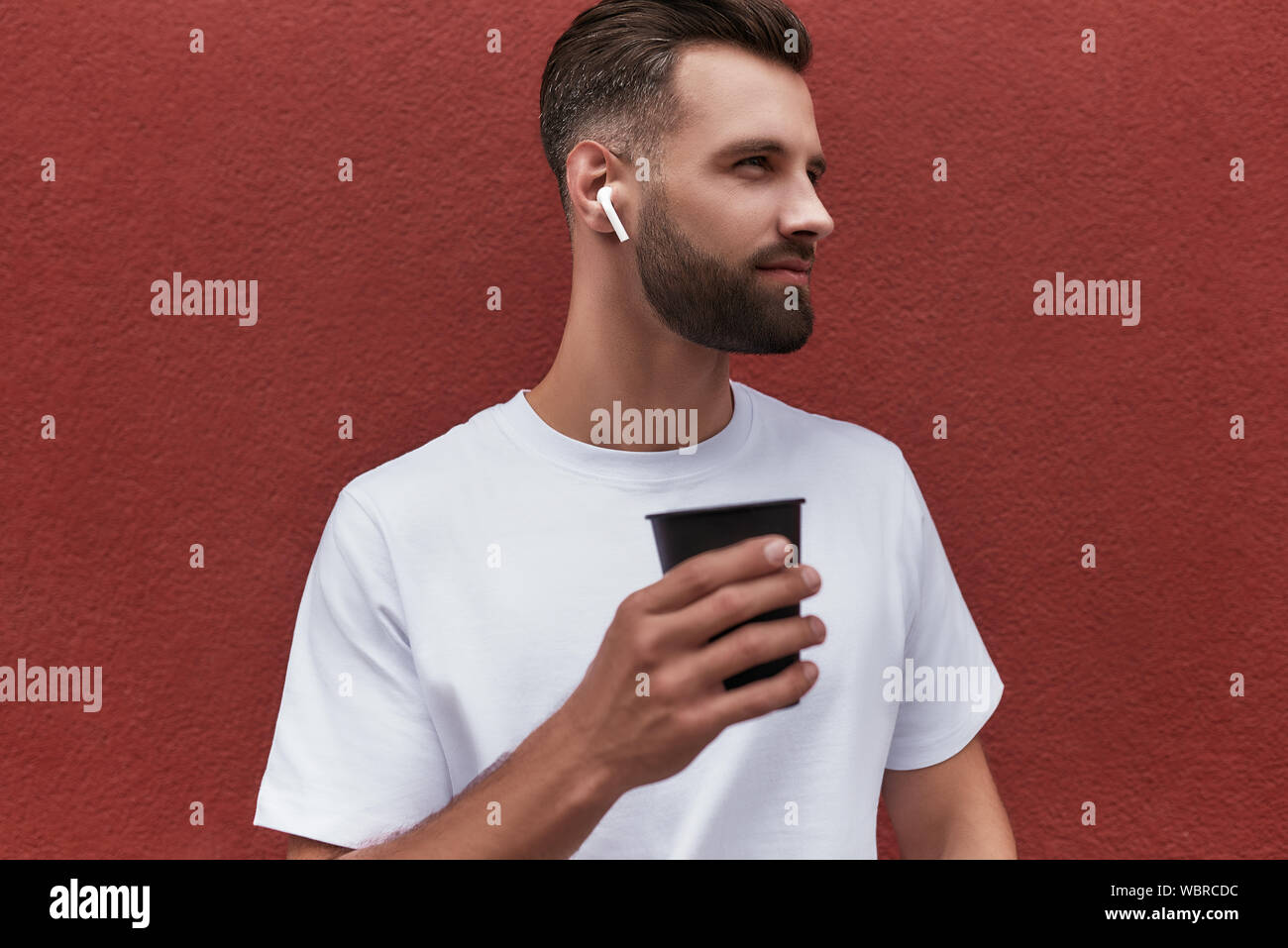 Gedanken über das Leben. Gut aussehender bärtiger Mann im Kopfhörer holding Tasse Kaffee und zur Seite schauen, während man gegen die Rote Wand im Freien. Das Leben in der Stadt. Männer Schönheit Stockfoto