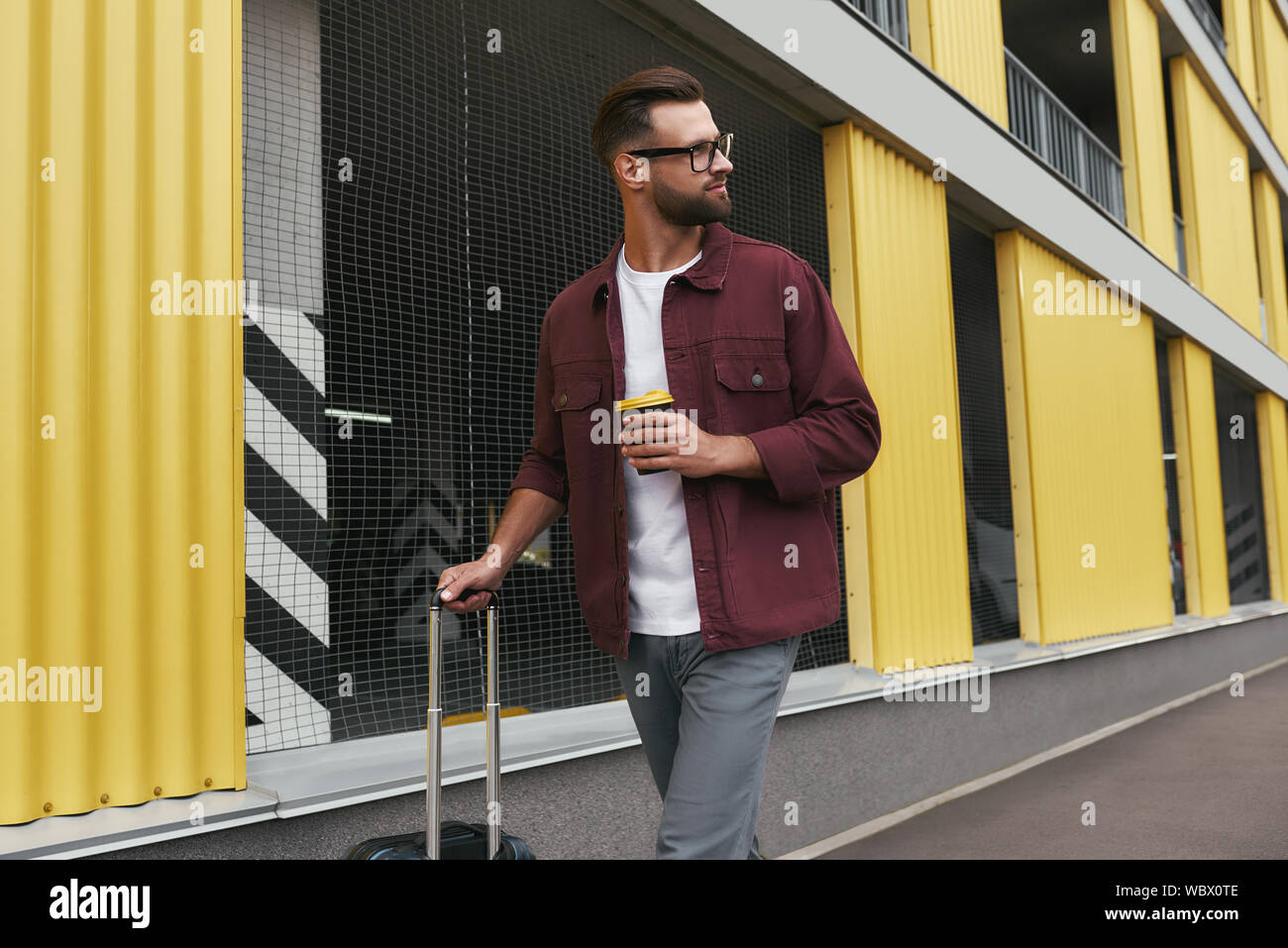 Tourist. Gut aussehender bärtiger Mann in Freizeitkleidung und Brillen mit einer Wegwerf- Cup und sein Gepäck beim Bummel durch die Stadt. Travel Concept. Reise Stockfoto