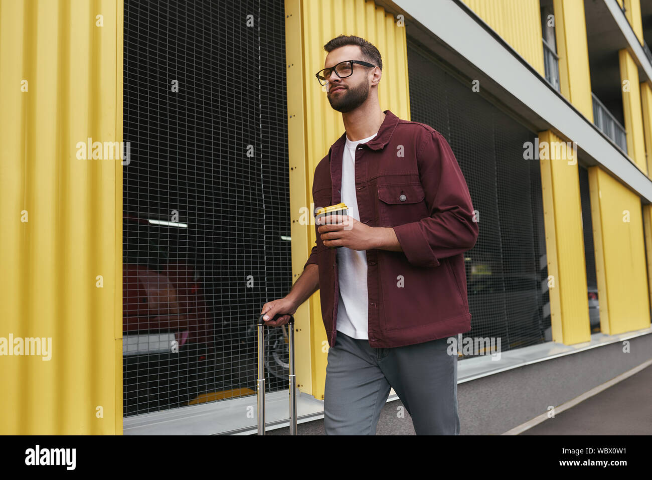 Die Zeit reisen. Schöner Lächeln bärtigen Mann in Freizeitkleidung und Brillen mit einer Wegwerf- Cup und sein Gepäck beim Bummel durch die Stadt. Travel Concept. Reise Stockfoto