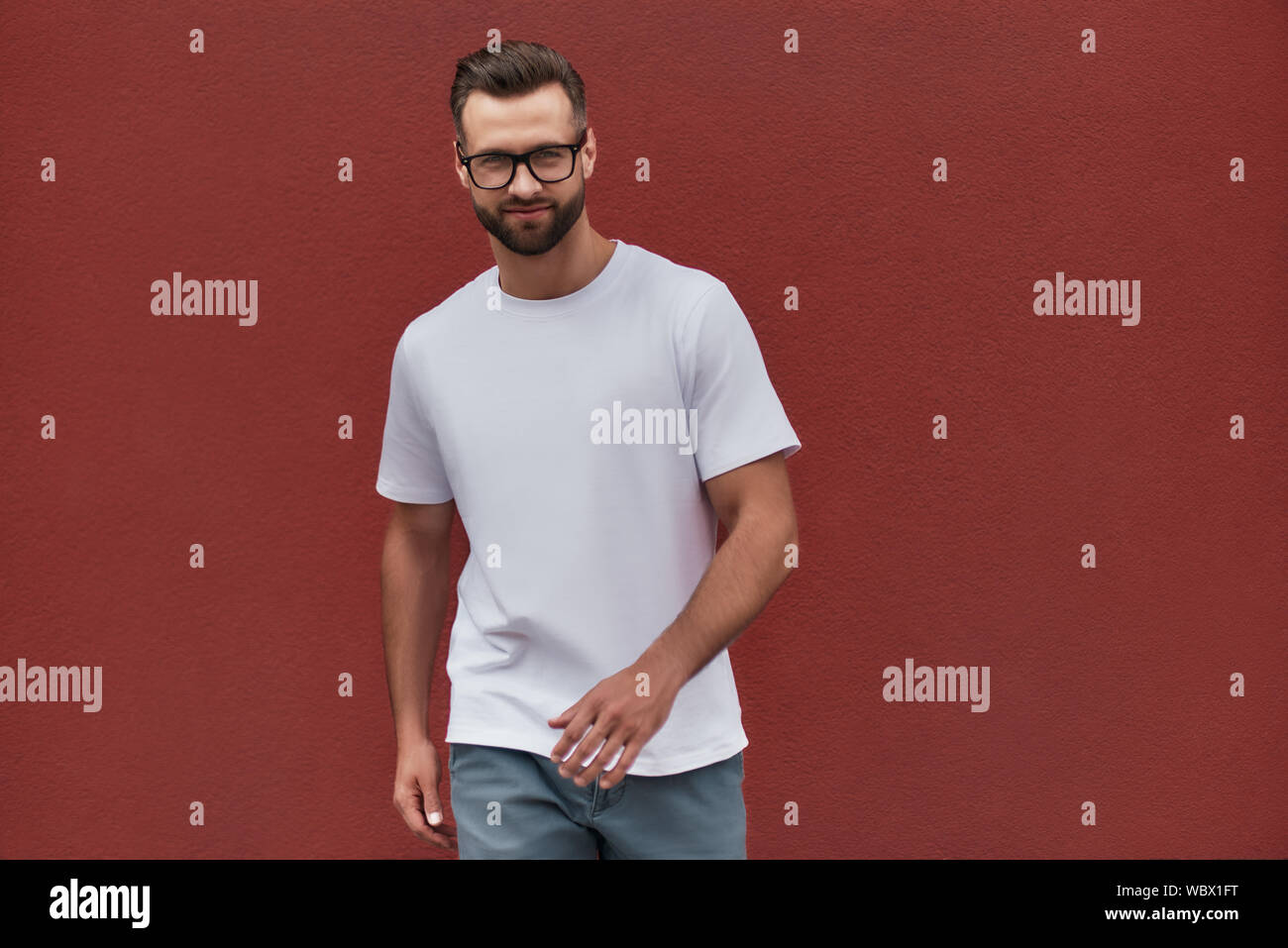 Jung und hübsch. Portrait von attraktiven bärtigen Mann in Brillen lächelnd und Kamera im Stehen gegen die Rote Wand im Freien. Männer Schönheit Stockfoto