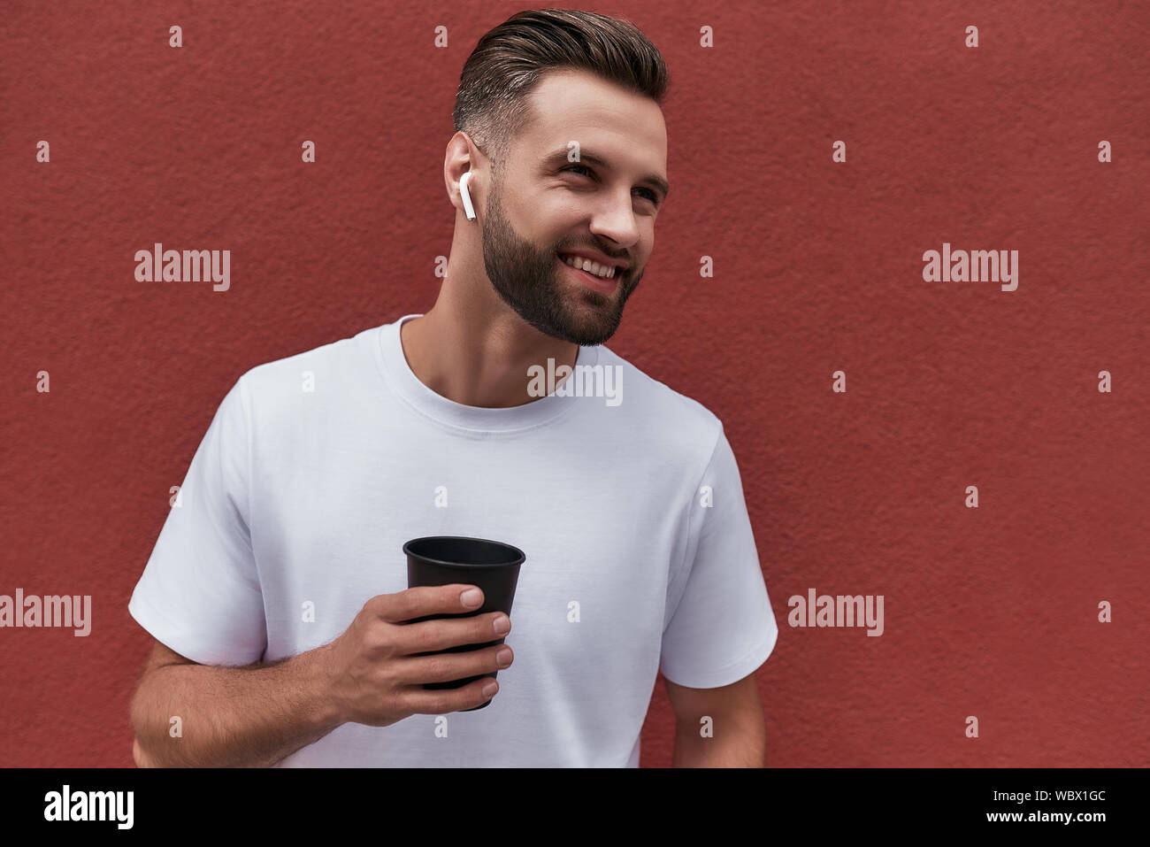 Jung und glücklich. Gut aussehender bärtiger Mann im Kopfhörer holding Tasse Kaffee und lächelnd, während man gegen die Rote Wand im Freien. Das Leben in der Stadt. Männer Schönheit Stockfoto