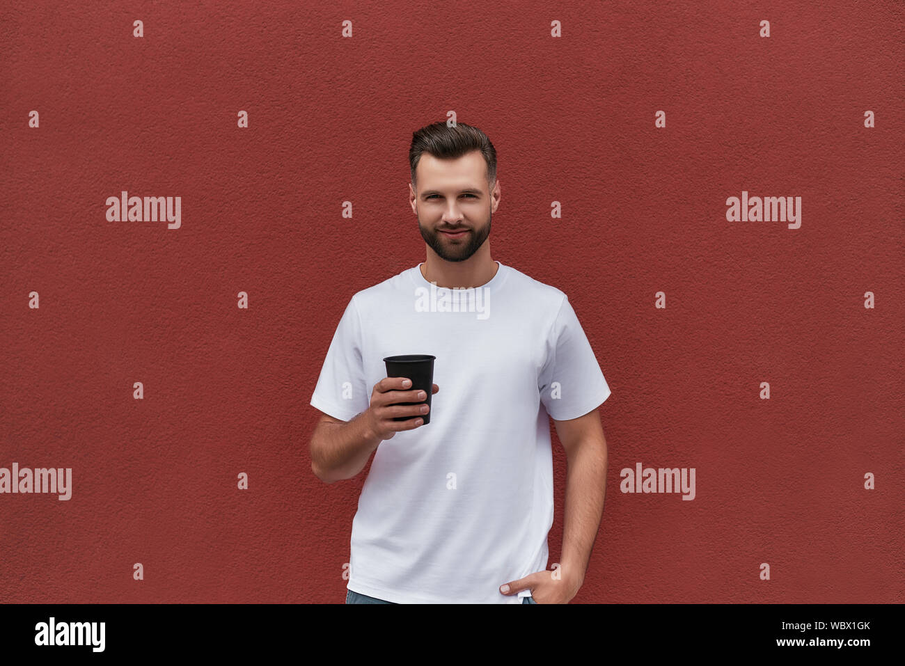 Gute Zeit. Schöner Mann in Freizeitkleidung holding Tasse Kaffee und Suchen an der Kamera, während gegen die Rote Wand im Freien stehend Stockfoto