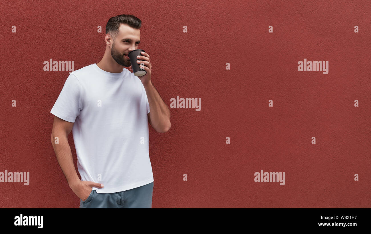Frischen Kaffee. Schöner Mann in Freizeitkleidung holding Tasse Kaffee und zur Seite schauen, während gegen die Rote Wand im Freien stehen. Web Banner Stockfoto
