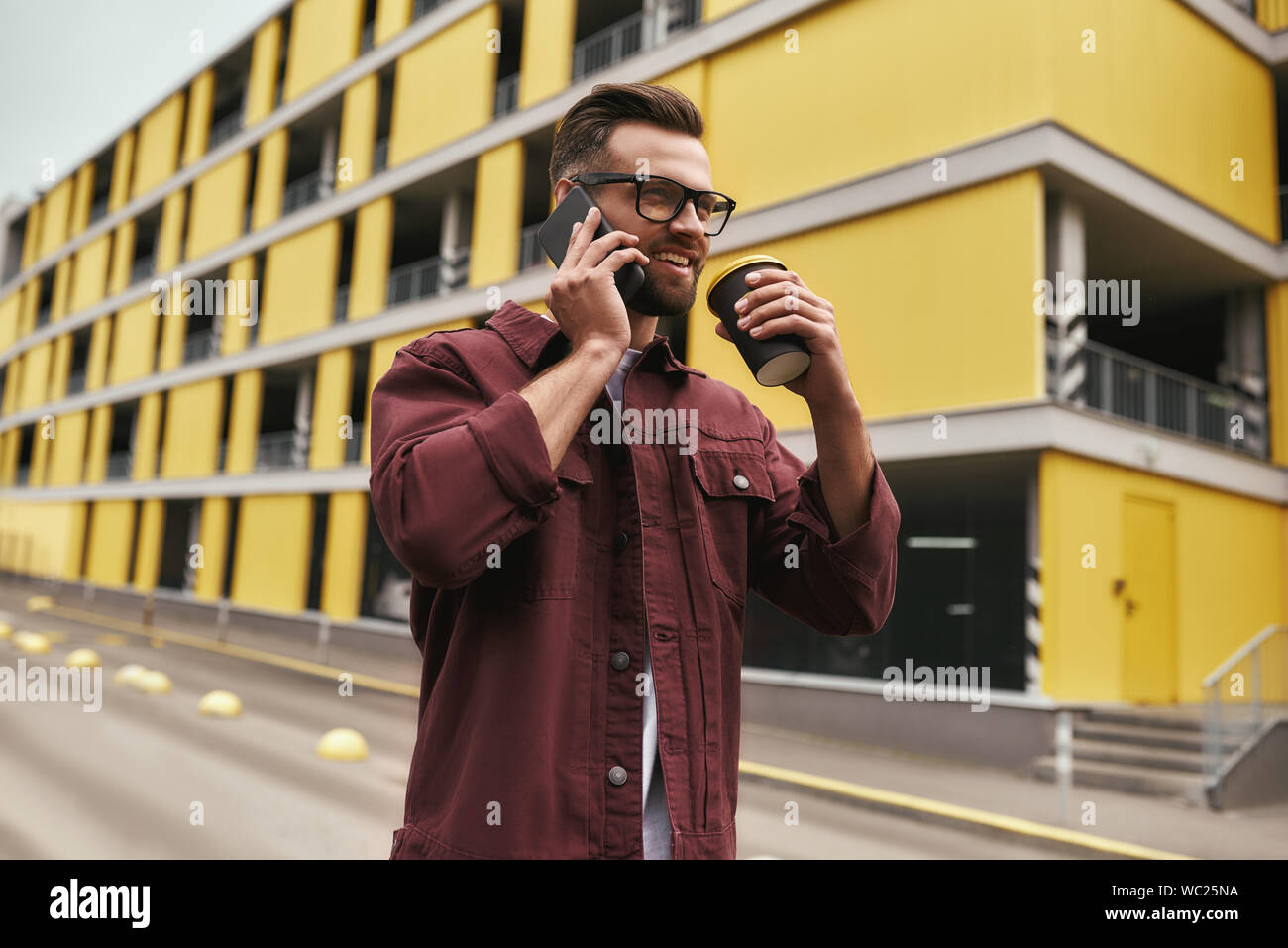 Große Nachrichten. Hübscher junger Mann in Freizeitkleidung und Brillen Kaffee trinken und reden über das Telefon mit jemandem beim Bummel durch die Stadt. Urban Style. Kommunikation Stockfoto
