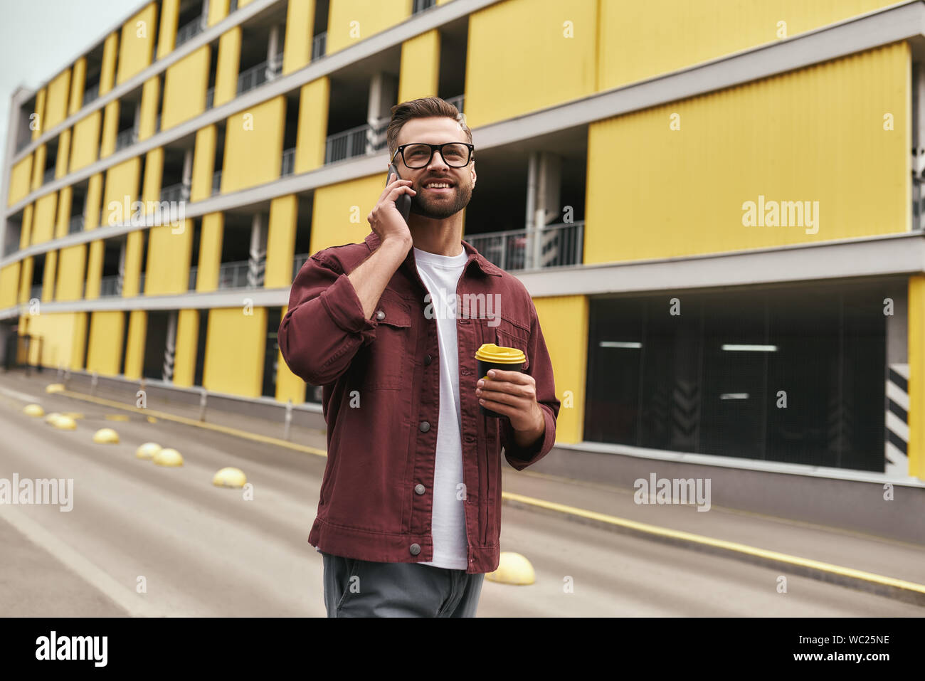 Glücklich, Sie zu hören. Hübscher junger Mann in Freizeitkleidung und Brillen mit einer Wegwerf- Cup und im Gespräch per Telefon mit jemandem beim Bummel durch die Stadt. Urban Style. Kommunikation Stockfoto