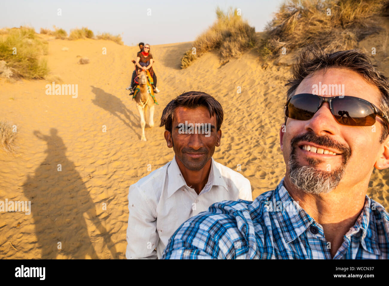 Ein kaukasischer Tourist auf einem Kamel Trek dauert eine selfie von sich selbst und seinem Fahrer sowie seine Frau auf ihrem Kamel mit ihrem Fahrer im Hintergrund. Stockfoto