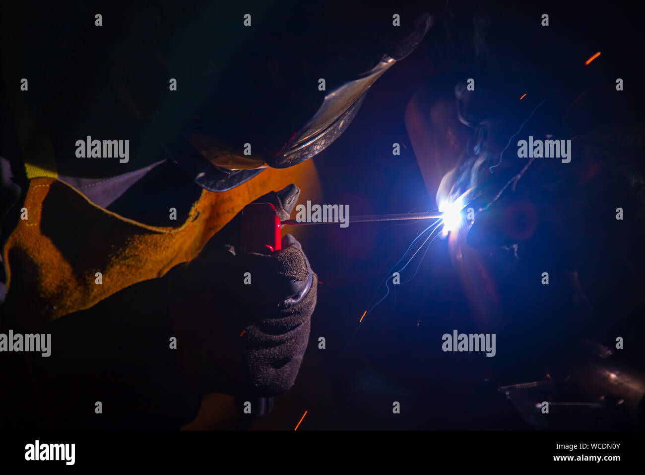 Ein Schweißer bei der Arbeit. Die Funken fliegen. Schweißer in der Maske bei der Arbeit. Beruf des Schweißers. Funken rund um. Schweißen von Metall. Der dunkle Hintergrund. Helles Licht. Stockfoto