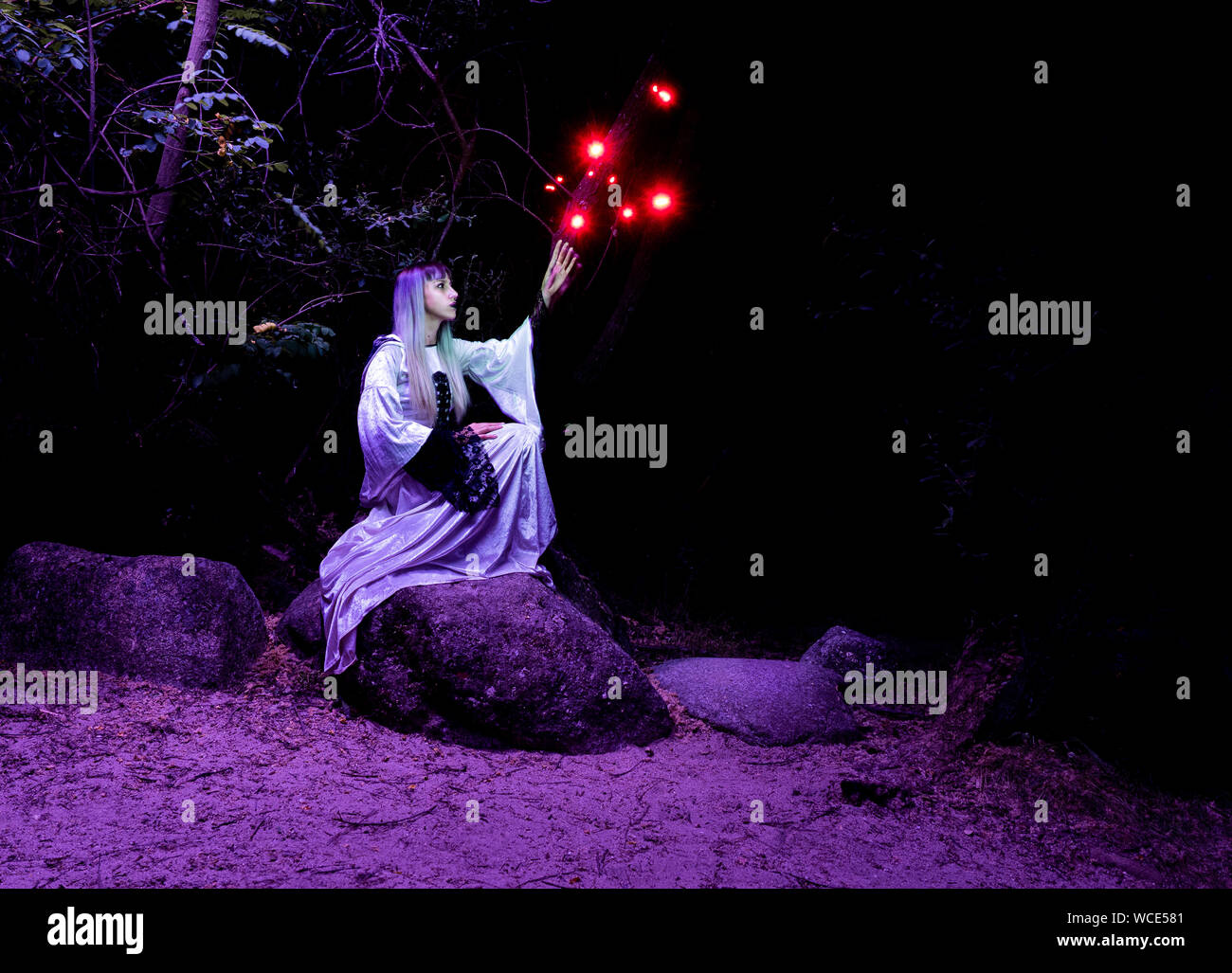 Eine Frau sitzt auf einem Felsen im Wald und in einer Tunika gekleidet, wirft die roten Lichter aus ihren Händen. Stockfoto