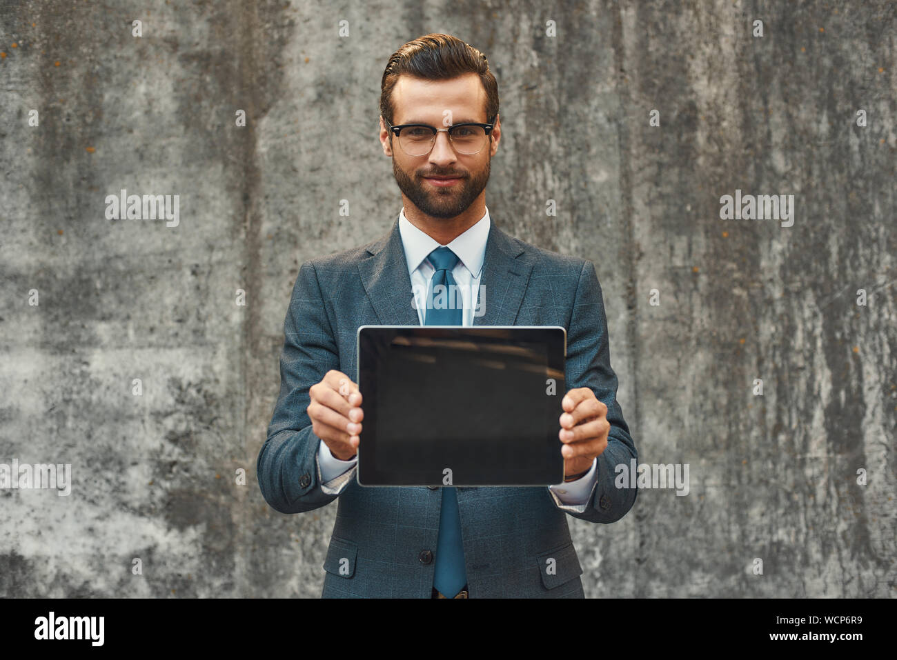Moderne Geschäftsmann. Portrait von gut aussehenden bärtigen Geschäftsmann in Brillen Übersicht digitale Tablet an der Kamera, während sich gegen grauen Stein Wand im Freien. Digitale Konzept. Business Konzept Stockfoto