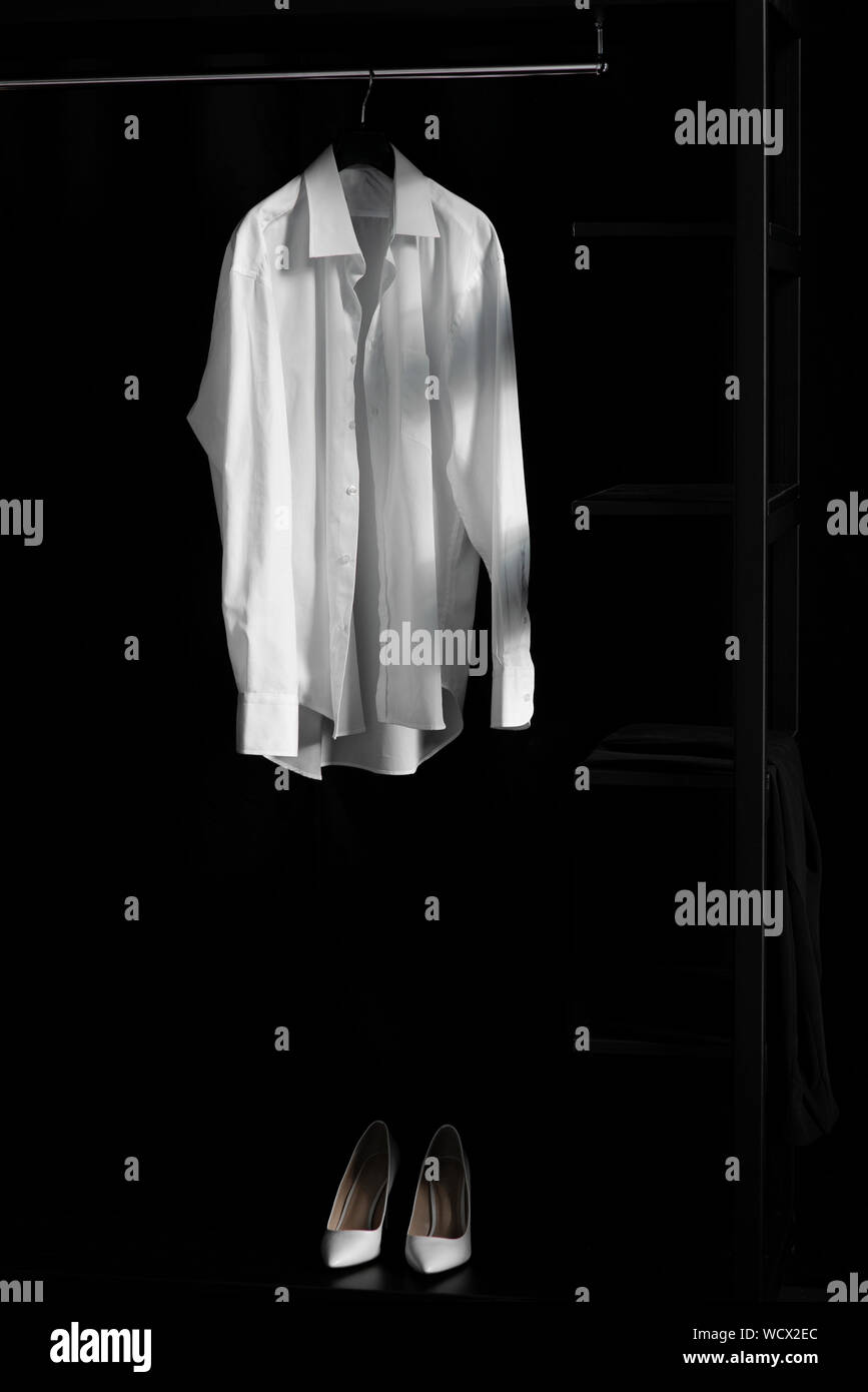 Weißes Hemd mit Bügel über Paar weiße Schuhe auf schwarzem Hintergrund. Fashion Concept Stockfoto