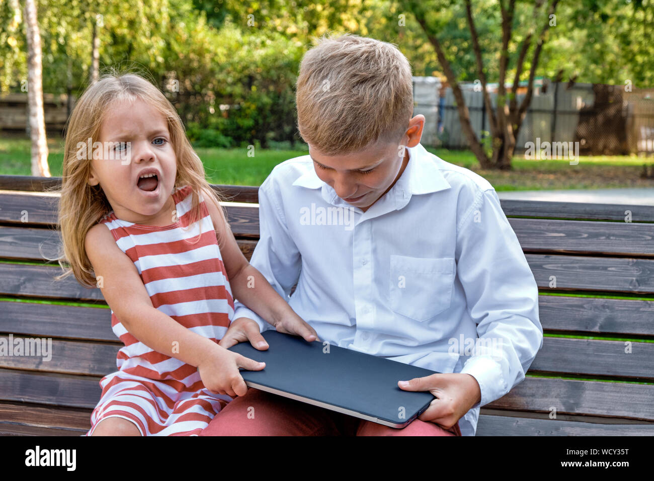 Konzept Gadget kämpfen. Ein Mädchen nimmt einen Laptop von einem Jungen auf einer Bank in einem Park. Stockfoto