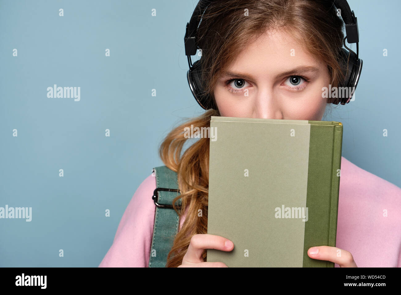 Ein junges Mädchen in einem rosa Pullover und Kopfhörer mit einem Rucksack steht auf einem blauen Hintergrund und blickt hinter einem Buch. Stockfoto