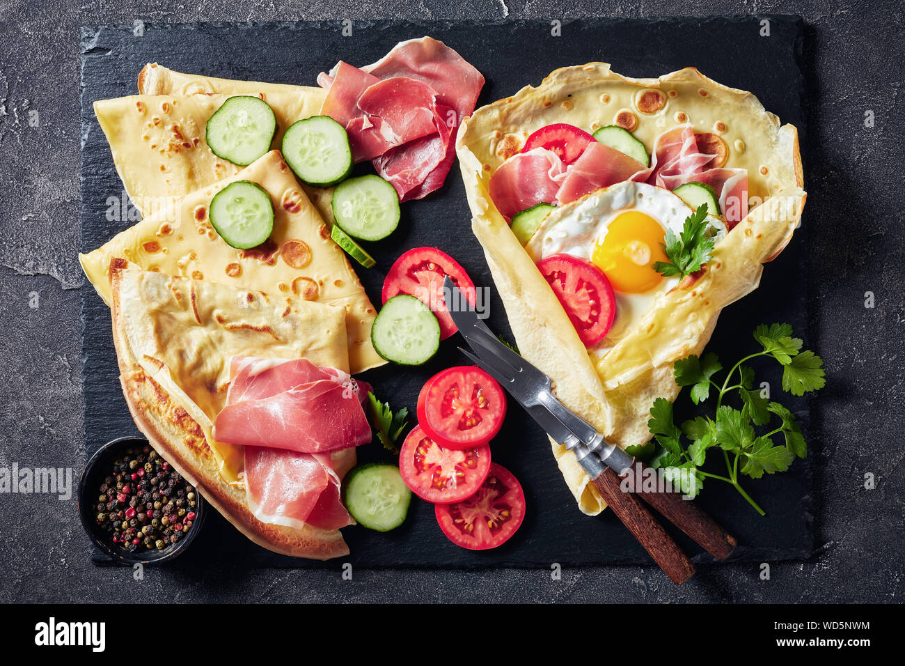 Köstliches Frühstück mit Crêpes mit Spiegelei, Jamon, Scheiben, frischen Tomaten und Gurken Füllungen auf schwarzem Schiefer Fach mit Zutaten, Ansicht von Abo Stockfoto