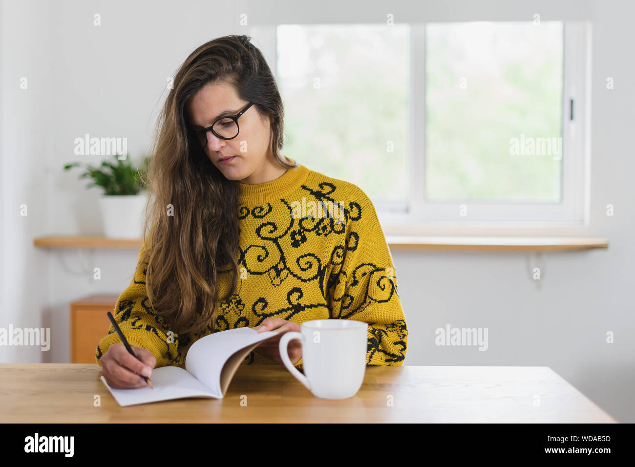 Frau mit Brille mit einem Bleistift in einem Notebook. Eine weiße Tasse auf einem hölzernen Werkbank in einem Haus. Stockfoto