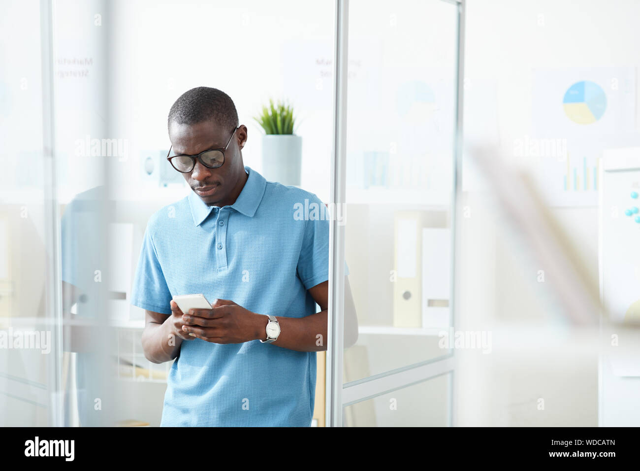 Junge afrikanische Büroangestellter in Freizeitkleidung an Büro- und Schreibarbeiten eine Nachricht auf dem Handy Stockfoto