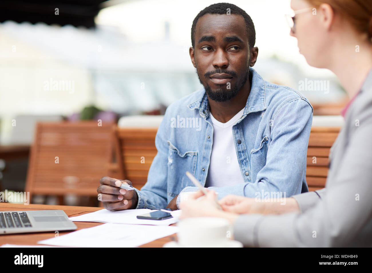 Afrikanische jungen Mann in Freizeitkleidung sitzen am Tisch und hören auf junge geschäftsfrau auf der Sitzung im Cafe Stockfoto