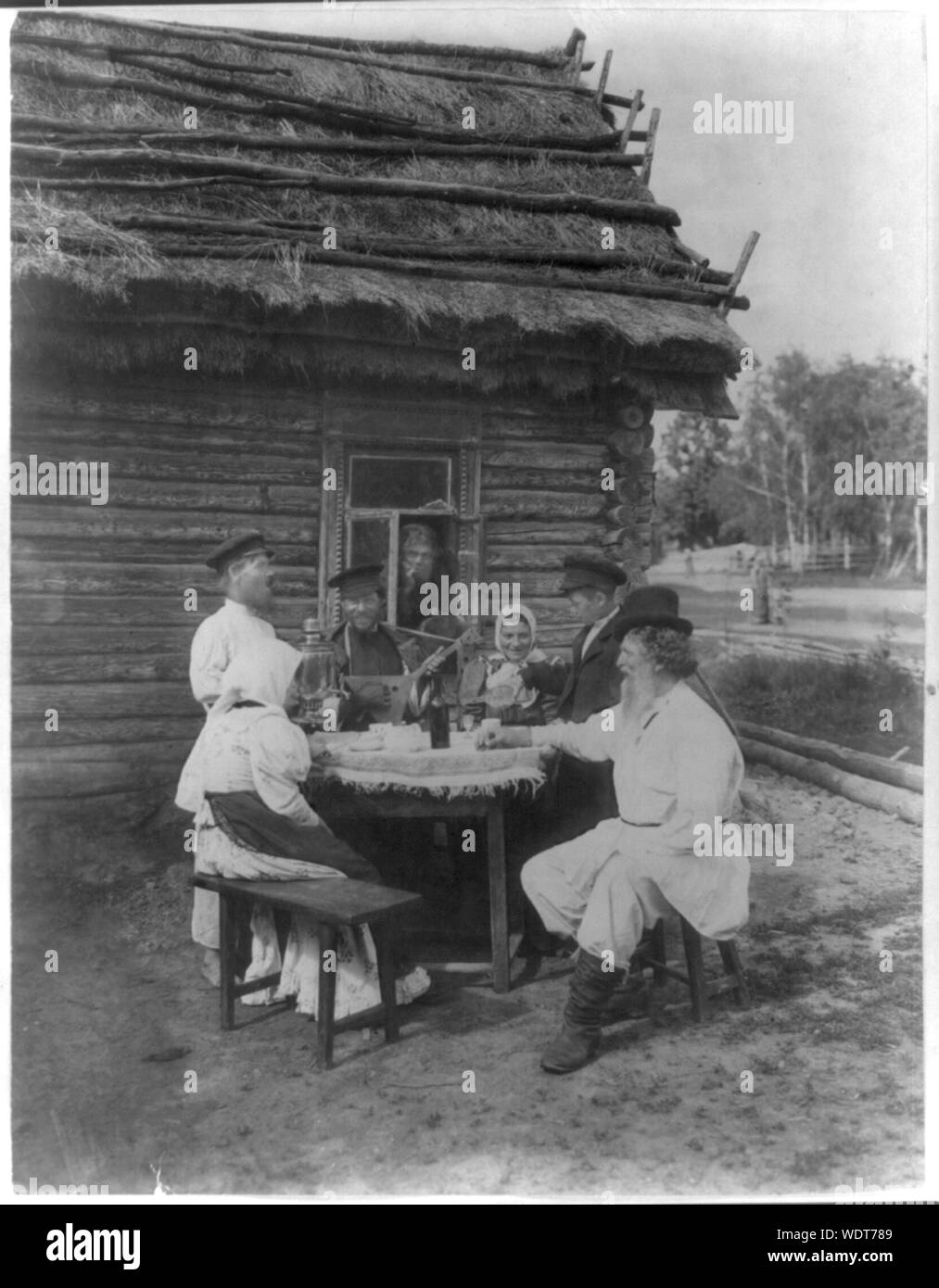 Gruppe der russischen Bauern stellte an einem Tisch im Freien Abstract: inszenierte genre Ansicht enthält Mann mit Balalaika, Samowar auf Tisch, junge Gießen trinken, man blickt durch Fenster und andere am Tisch draußen sitzt einem strohgedeckten Haus anmelden. Stockfoto