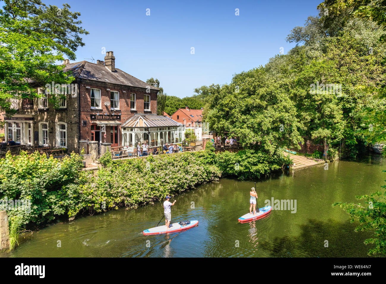 29. Juni 2019: Norwich, Norfolk, Großbritannien - Paar Paddle Boarding am Fluss Wensum, mit dem Red Lion Hotel im Hintergrund. Hellen Sommertag, viel Stockfoto