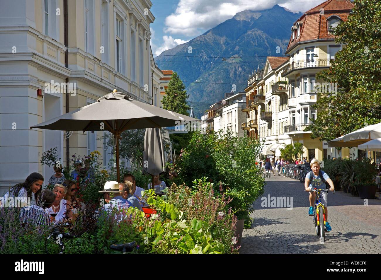 Italien, Autonome Provinz Bozen, Meran, Kind Reiten Fahrräder und Menschen auf der Terrasse eines Cafés in der Innenstadt sitzen mit den grünen Bergen im Hintergrund Stockfoto