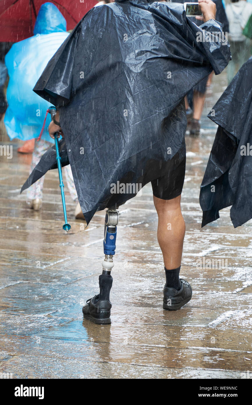 Aktive männliche Touristen oder Pilger mit Prothese zu Fuß auf einer Straße der Altstadt von Santiago de Compostela an einem regnerischen Tag. Positive behinderten Person Stockfoto