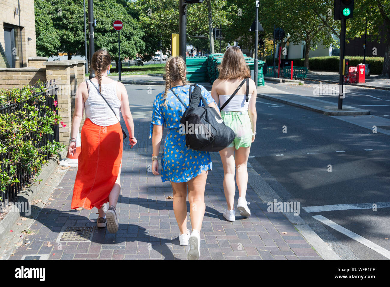 Drei junge Frauen, die in sommerliche Kleidung wandern mit dem Rücken zur Kamera entlang einer Straße im Südwesten von London, Großbritannien Stockfoto