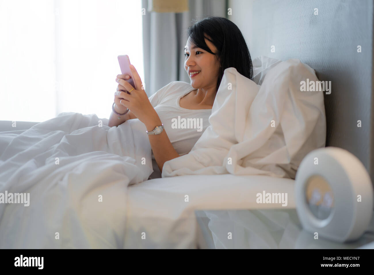 Morgen mit Gadget. Asiatische schöne junge Frau auf dem Bett liegend mit Smartphone. Stockfoto