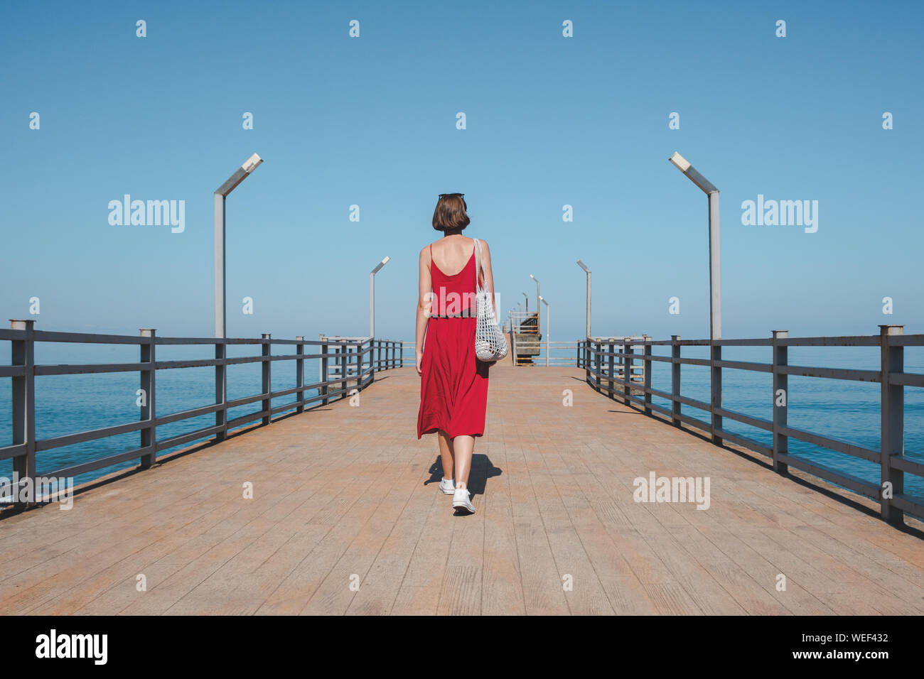 Wandern entlang der Pier mit dem blauen Meer. Die Frau im roten Kleid geht entlang der Kais. Schuß von hinten Stockfoto