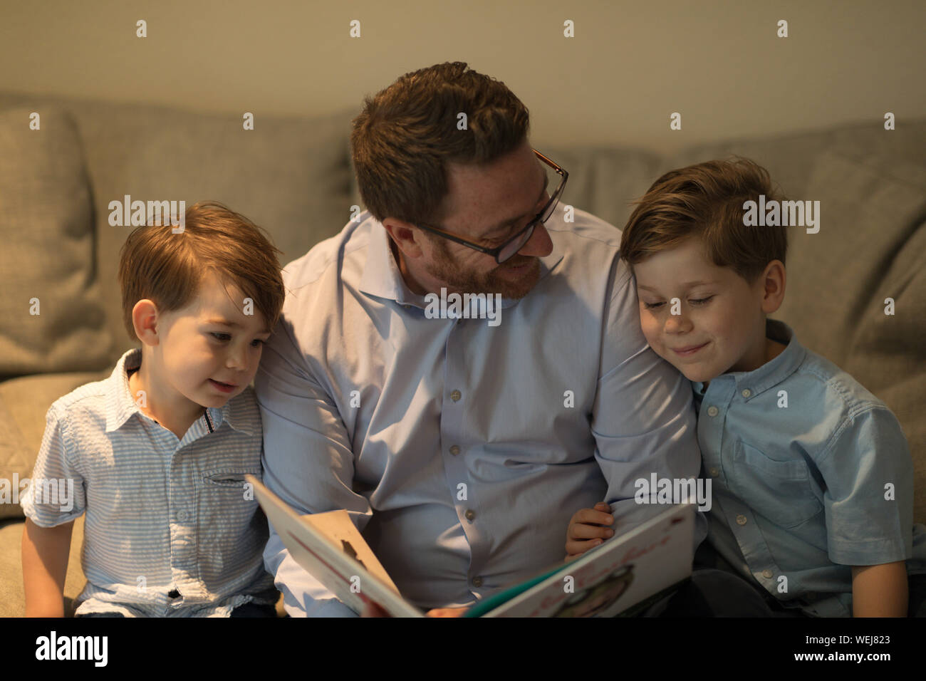 Vater mit zwei Söhnen, 2 und 4, auf der Suche bei Buch auf dem Sofa Stockfoto