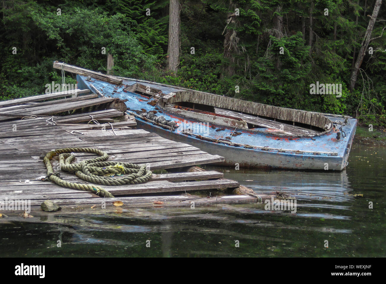 Ein verfallenes, stripped down Alte dayboat in den Untiefen zu einem baufälligen Dock sitzt auf einem bewaldeten Ufer in einer ruhigen Gegend an der Küste von Britisch-Kolumbien. Stockfoto