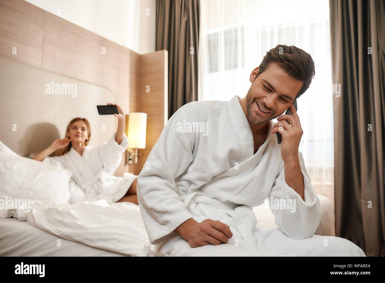 Portrait von Paar tragen weiße Bademäntel. Mann sitzt auf der Kante des Bettes und Gespräche am Telefon, während Frau nimmt selfie. Zusammen reisen Konzept. Horizontale Schuß Stockfoto