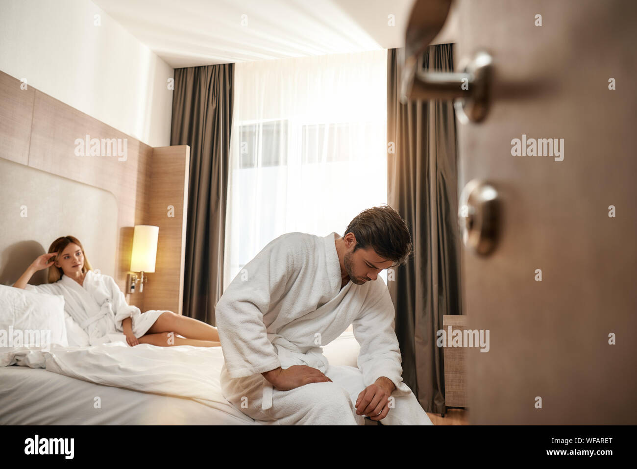 Portrait von Paar tragen weiße Bademäntel. Mann sitzt auf der Kante des Bettes, während Frau an ihm ist. Zusammen reisen Konzept. Horizontale Schuß Stockfoto