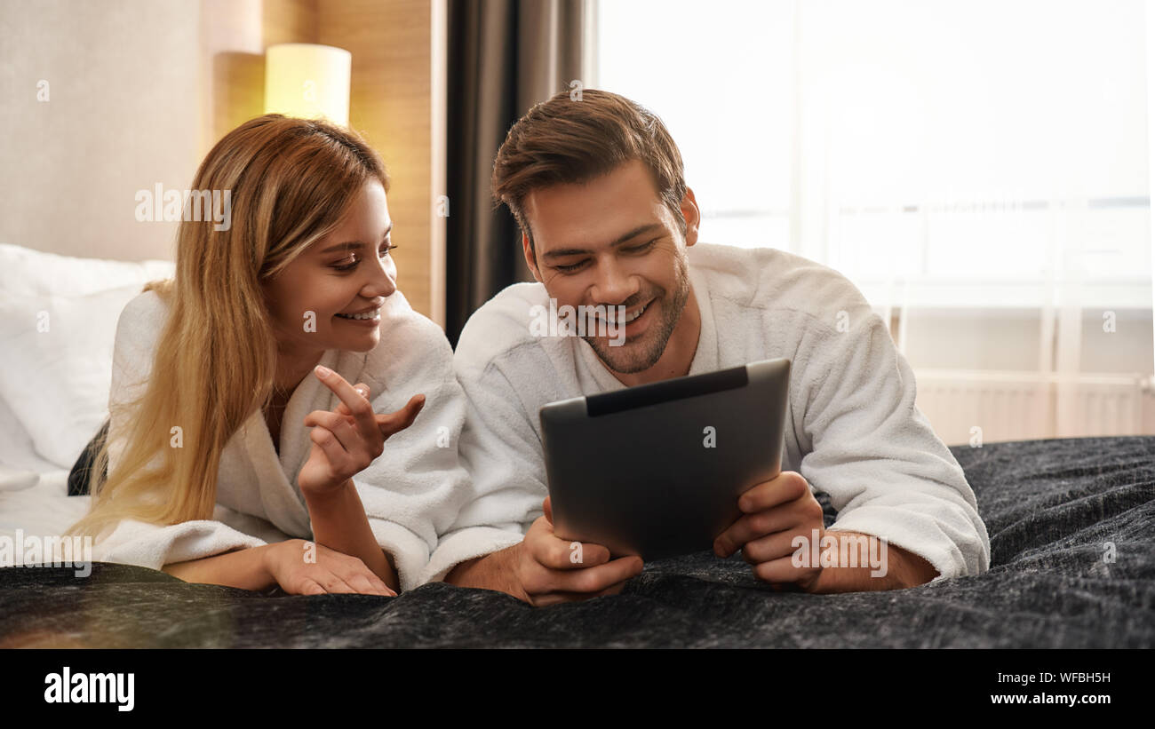 Portrait von Paar tragen weiße Bademäntel liegen auf dem Bett und auf den Bildschirm des Tablet-PC. Zusammen reisen Konzept. Horizontale erschossen. Ansicht von vorn Stockfoto