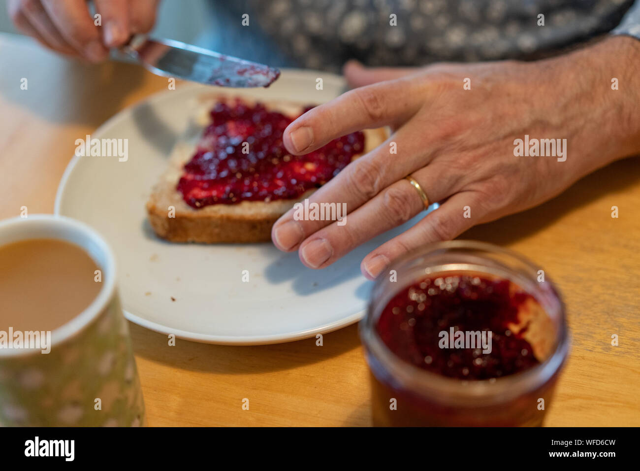 Ein Mann in der Küche sitzt und Verbreitung Gelee auf sein Brot mit Kaffee und ein Glas Gelee auf die Birke Tabelle. Stockfoto