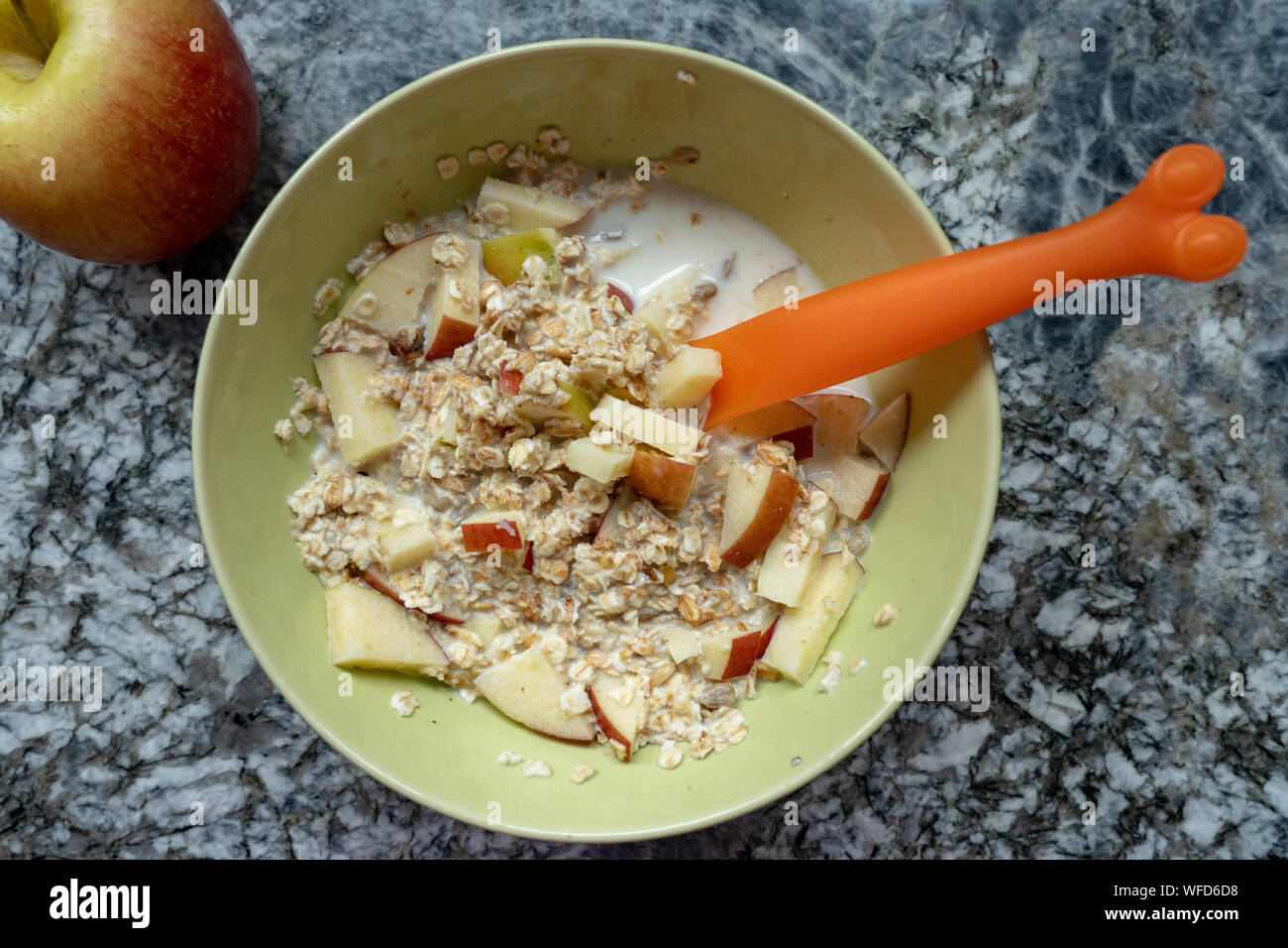 Vorbereitung eines Schweizer Müsli mit frischer Milch, Getreide, trockenen Trauben und Äpfel in einem hellgrünen Schüssel und eine orange Löffel auf einer Plattform aus Granit in der Küche Stockfoto