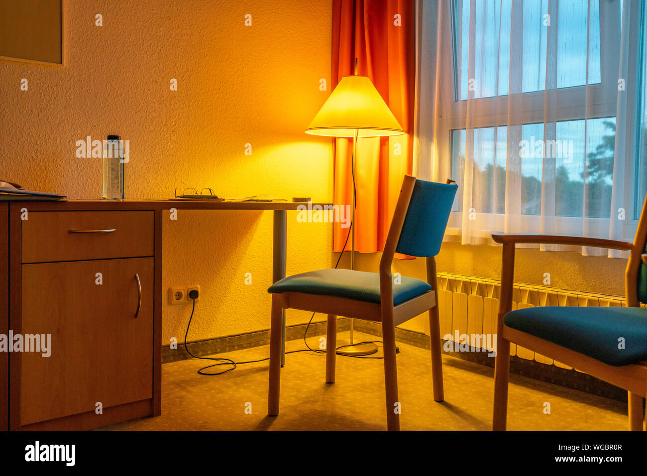 Ein Zimmer mit goldenen Licht von gelb Stehleuchte gefüllt mit zwei blauen Stühlen und ein Fenster mit orangefarbenen Vorhängen Stockfoto