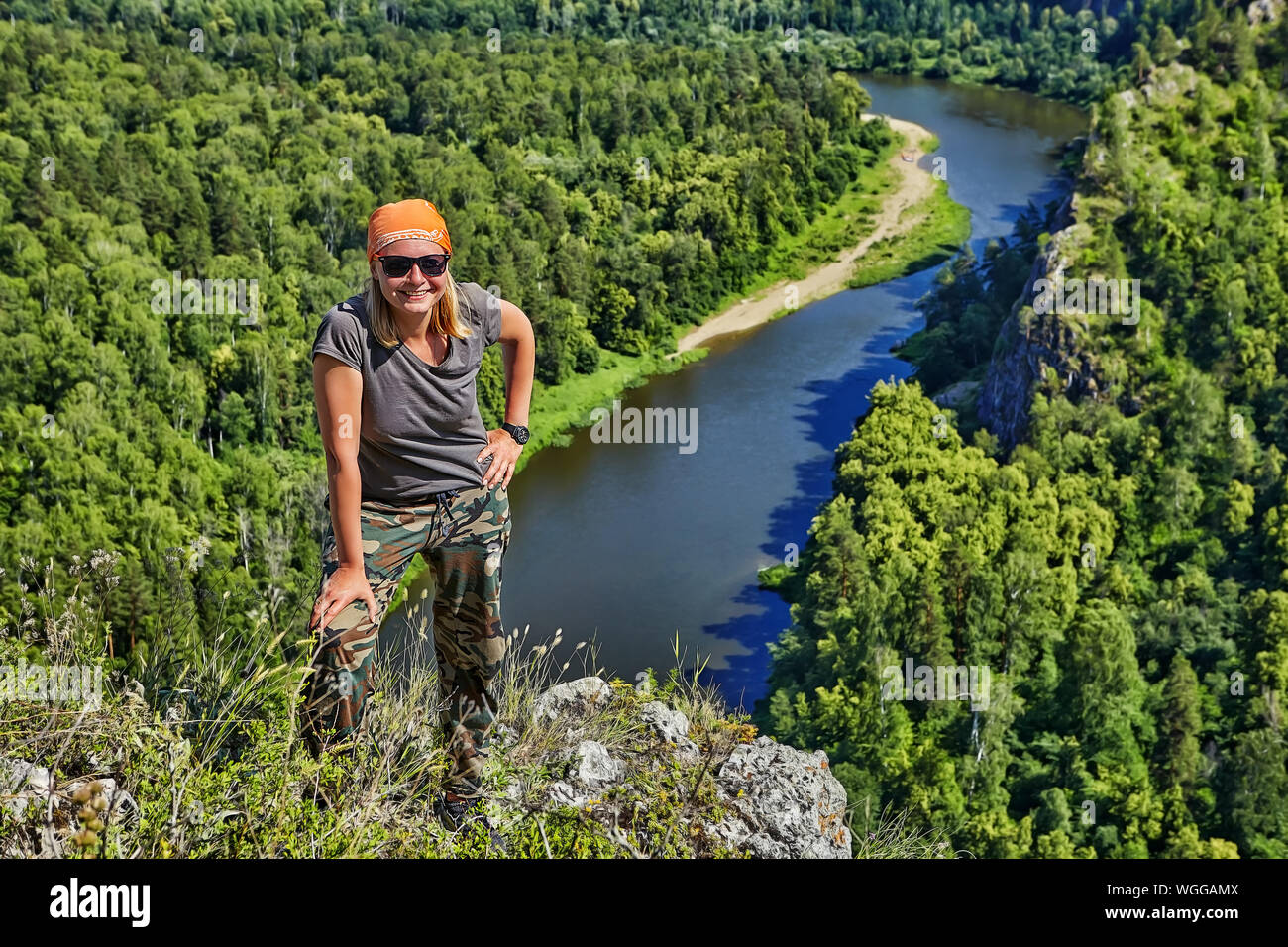Eine lächelnde Frau in einem bandana und Sonnenbrille steht auf einem Berg, vor dem Hintergrund einer ruhigen Wald Fluss unten, ein Wanderer ist enga Stockfoto