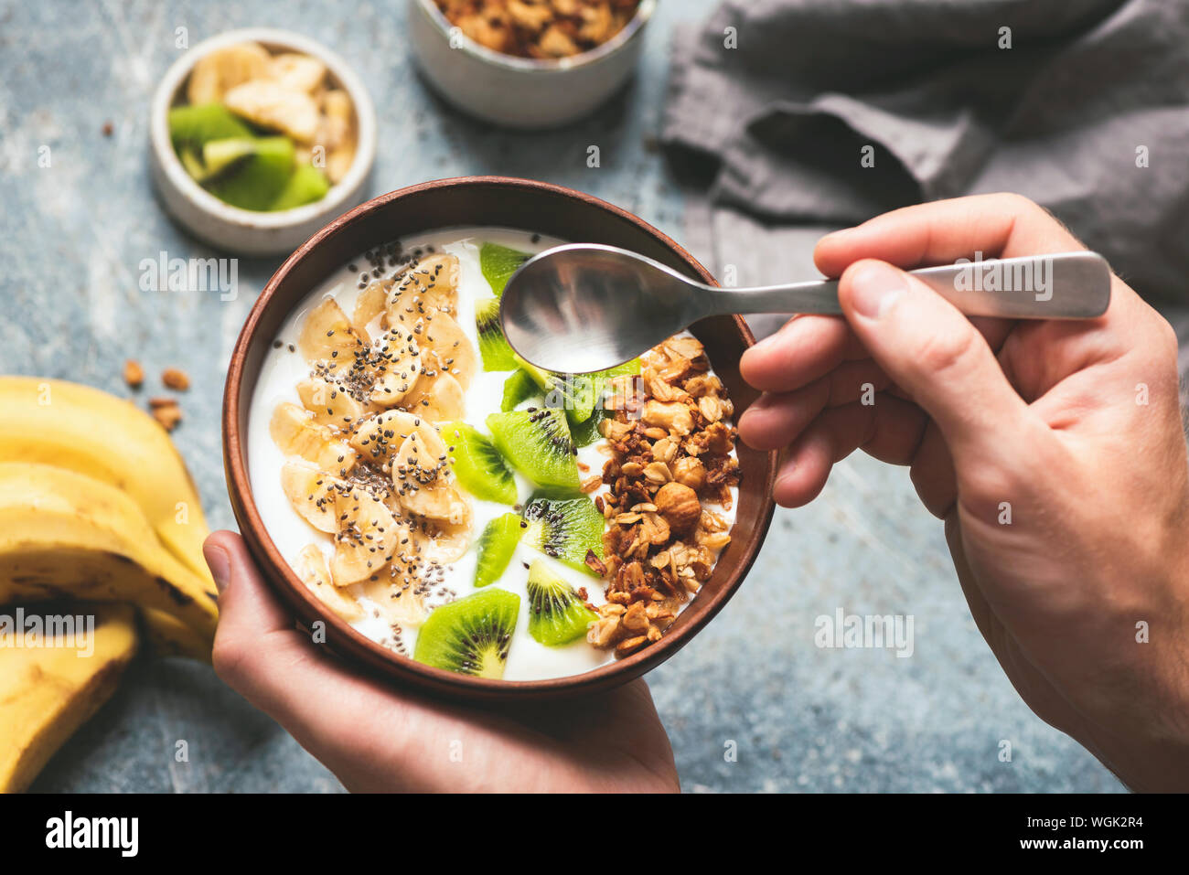 Gesund essen Frühstück Joghurt Schale mit Müsli und Obst Kiwi, Bananen, Kokosnuss. Männliche Hände halten Kokosnuss Schale mit Joghurt und Früchte Stockfoto