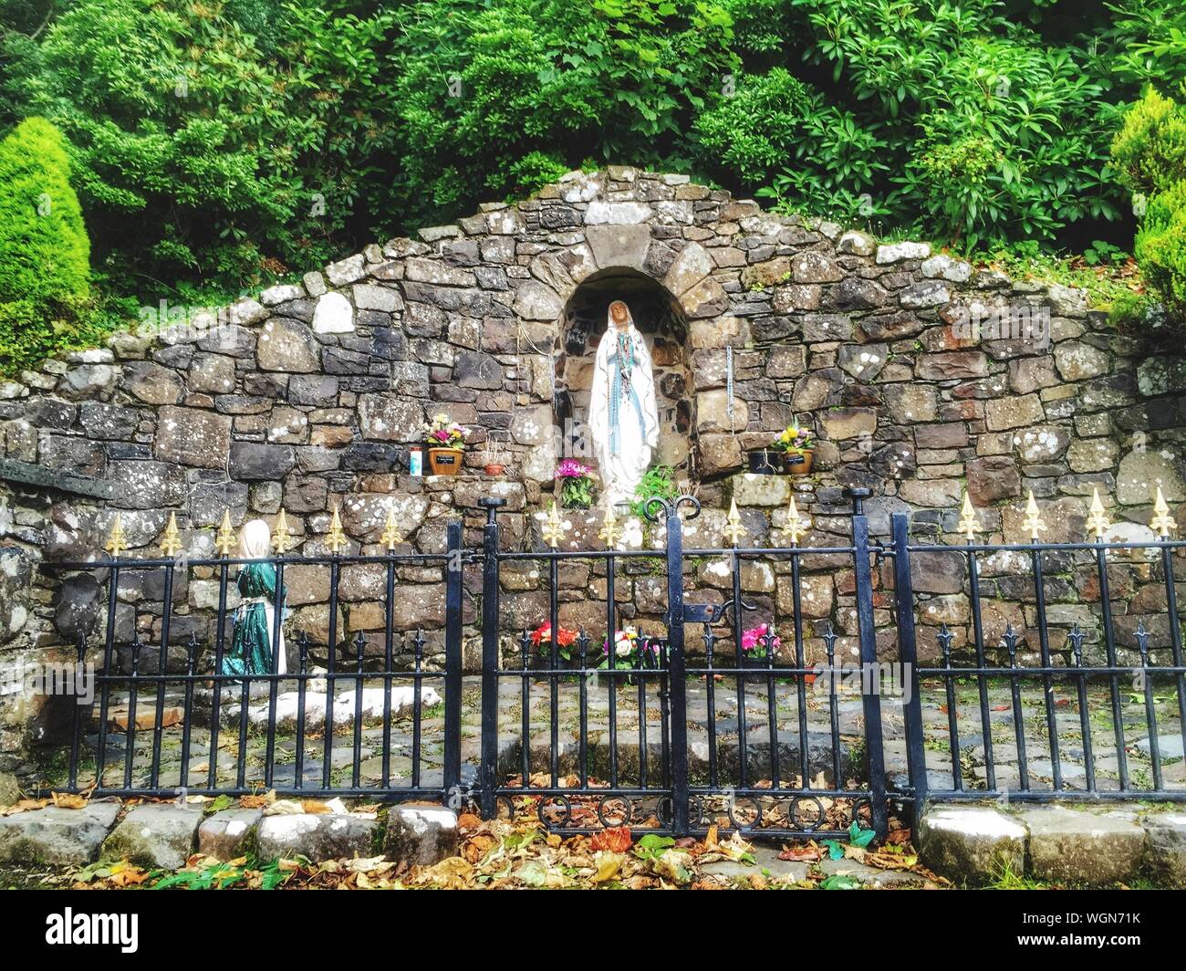 Jungfrau Maria Statue In Grotte vor Metallzaun gegen grüne Bäume Stockfoto