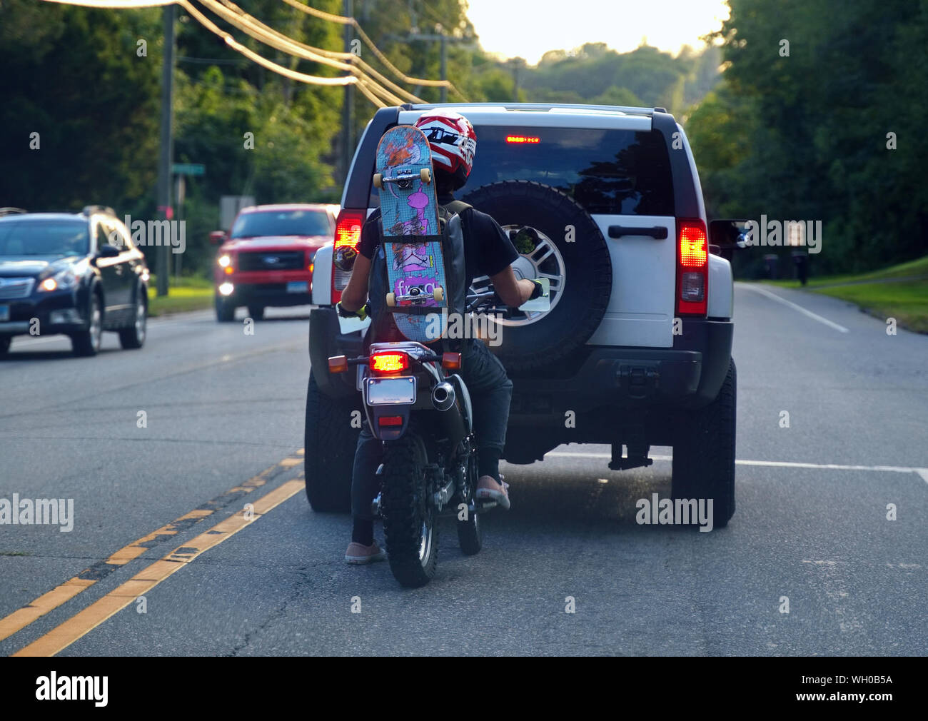 Storrs, CT USA. Aug 2019. Junge Motorradfahrer an einer Kreuzung mit einem Skateboard festgeschnallt auf dem Weg zum Skatepark zu Rucksack. Stockfoto