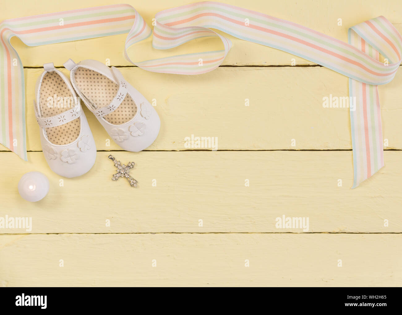 Taufe Hintergrund mit weißen Schuhen, Band, Kerze und Kristall Kreuz Anhänger auf blass gelb lackierten Holz- Hintergrund - Ansicht von oben Foto Stockfoto