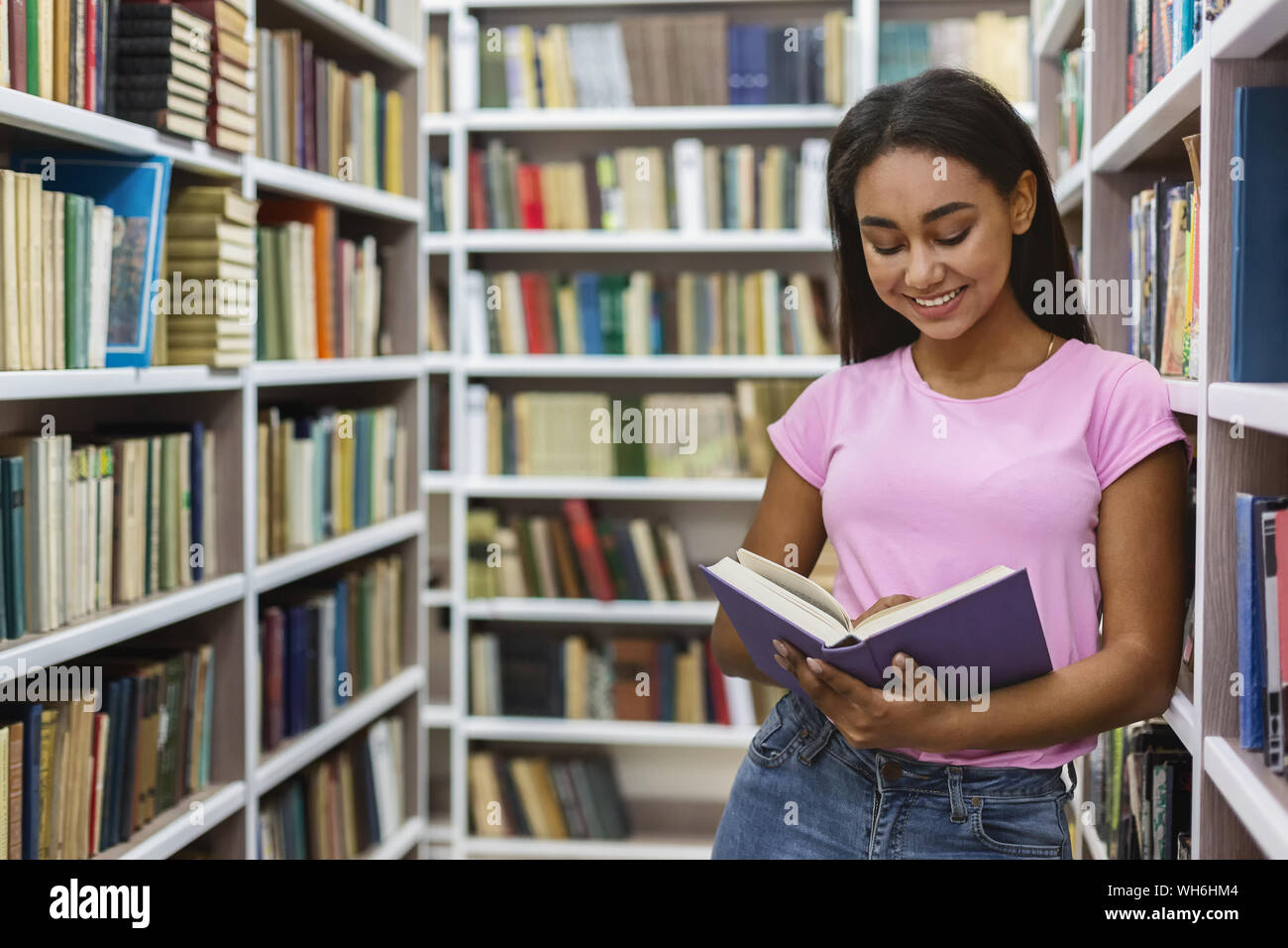Lächelnd schwarze Mädchen lesen Buch zwischen Bücherregalen Stockfoto