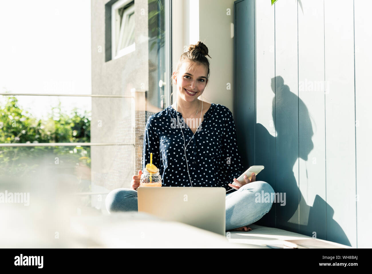 Lächelnde junge Frau mit Laptop, Handy und gesundes Getränk zu Hause Stockfoto